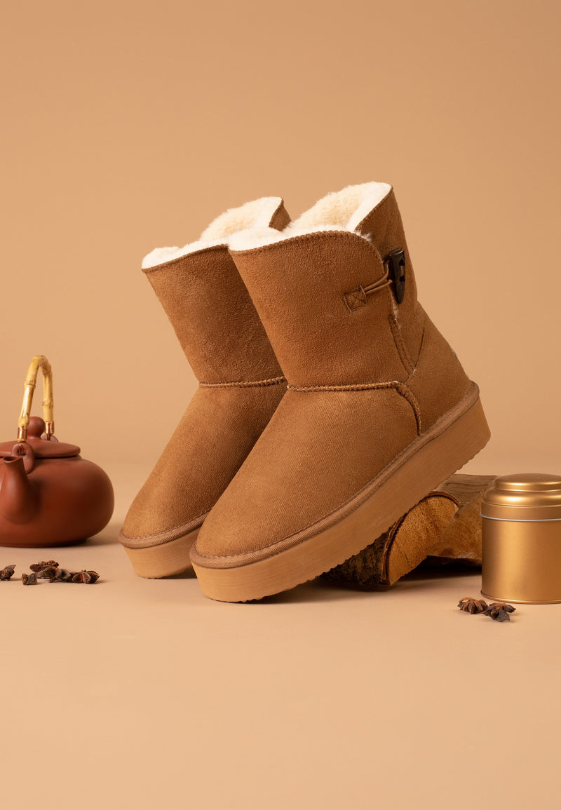 Paire de bottines fourrées Anais camel posées sur fond caramel, pot doré à côté