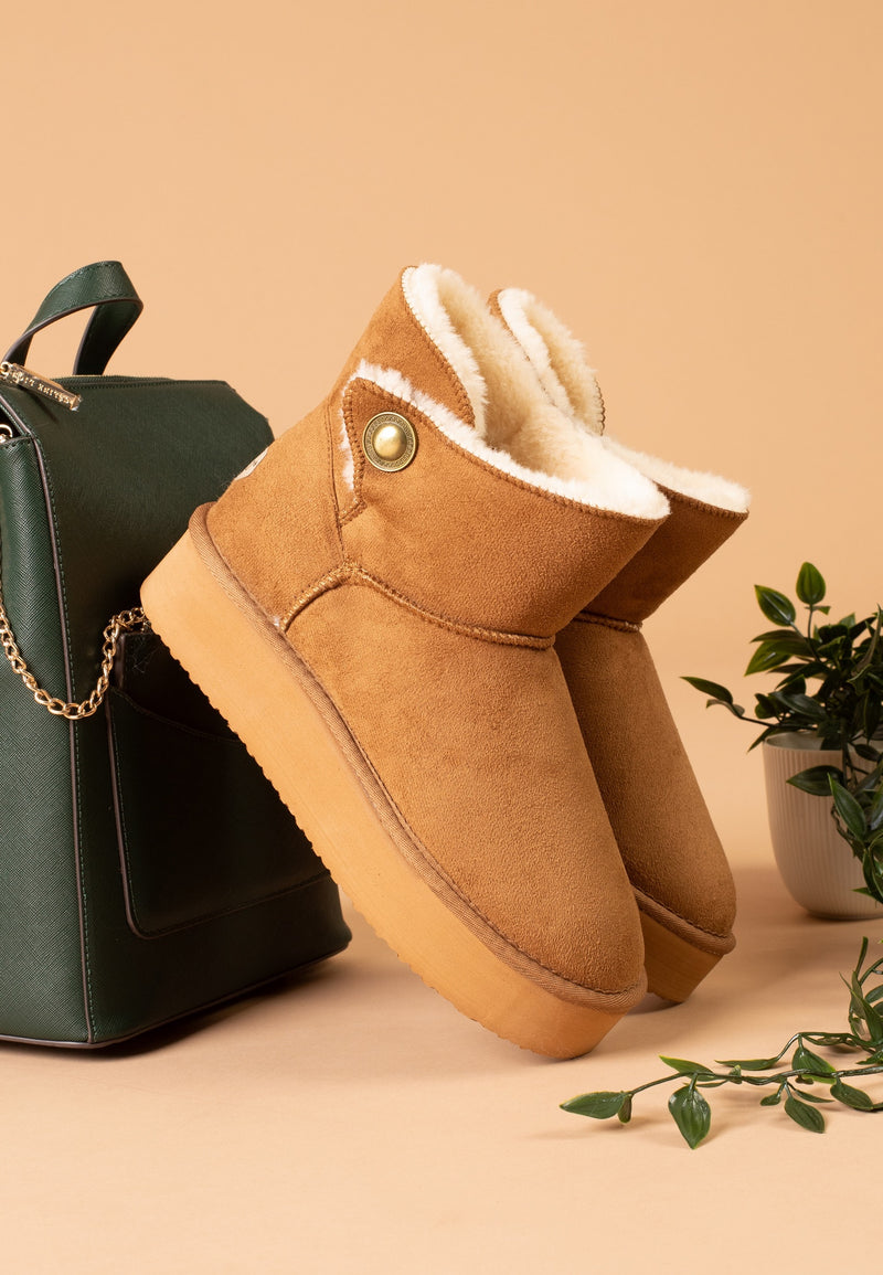 Bottines fourrées Cayuga camel de trois-quarts, posées contre un sac à dos vert foncé
