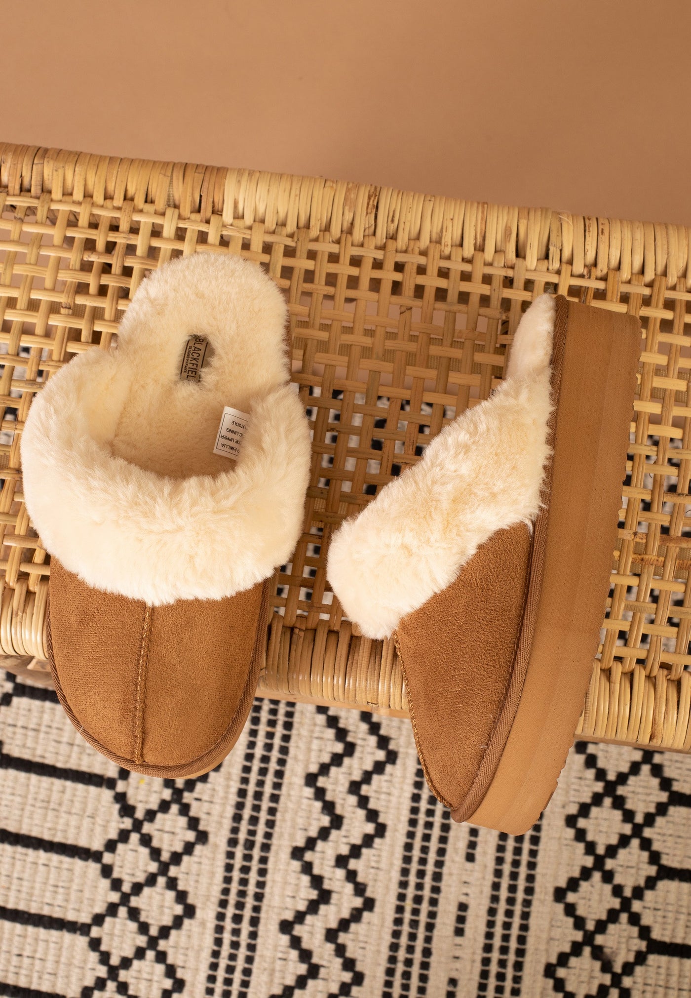 Sous une chaise en cannage, la paire de chaussons fourrés camel Sleepers Emilija