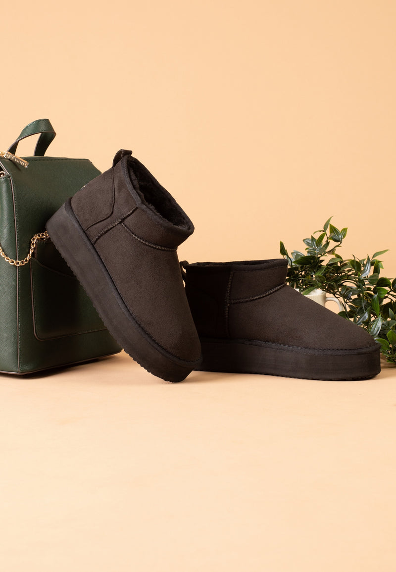 Bottines fourrées Milli noires posées de profil près d'un sac kaki et d'un feuillage