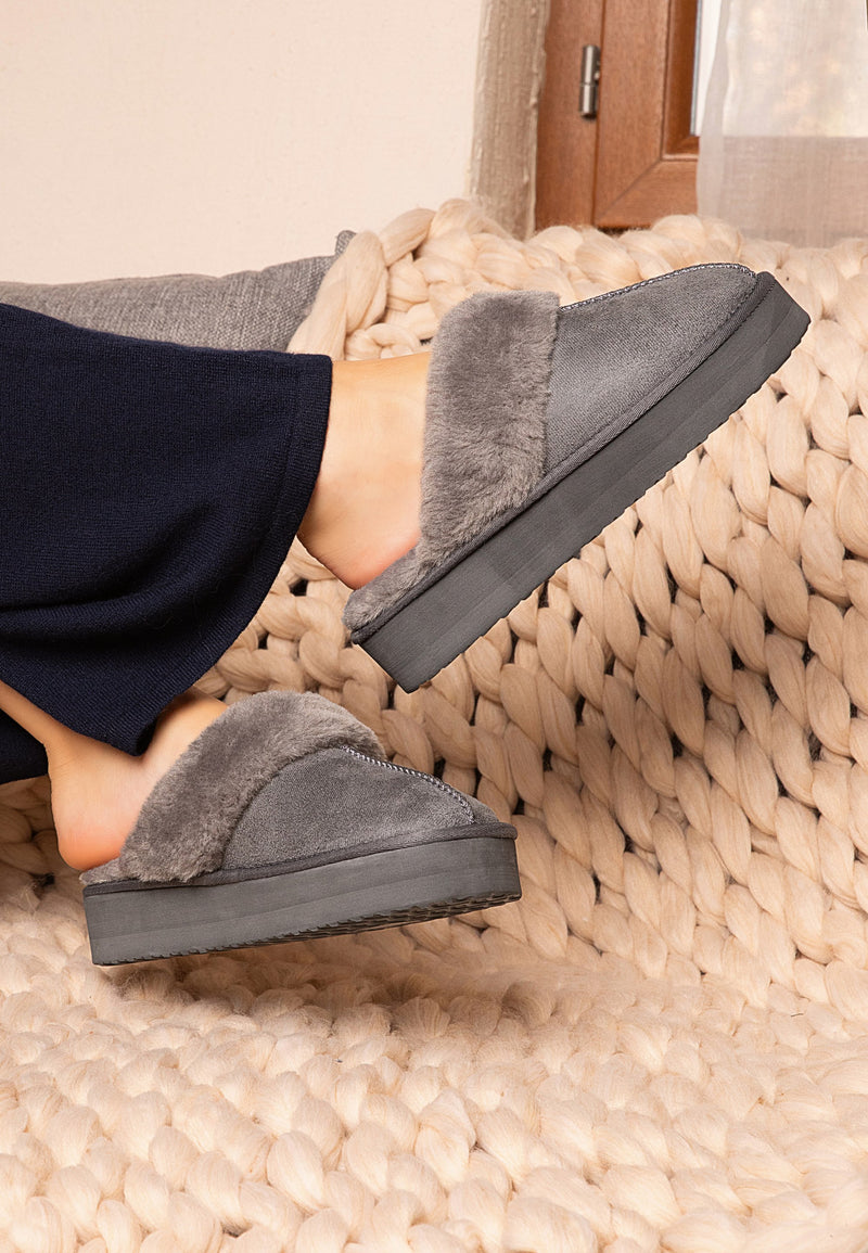 Chaussons fourrés Emilija anthracite portés sur un plaid en grosse maille, bas marine