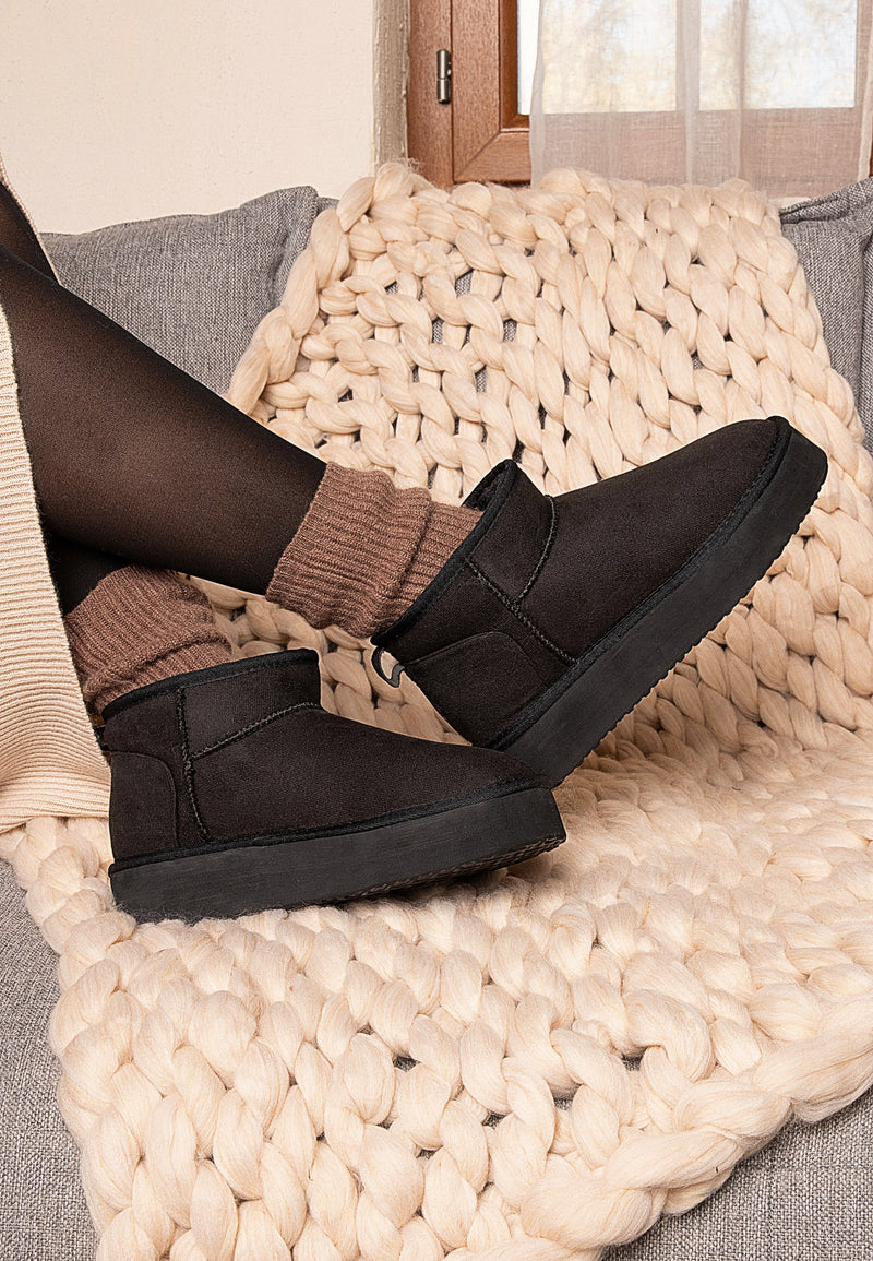 Pieds chaussés de bottines fourrées Milli noires, portées sur un plaid écru