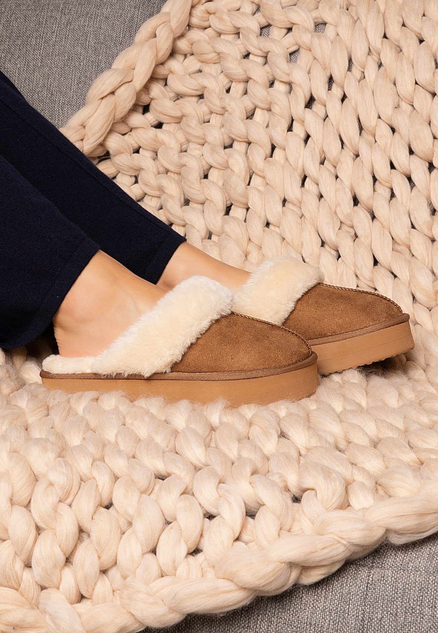 Sur un plaid en grosse maille, la paire de chaussons fourrés camel Sleepers Emilija