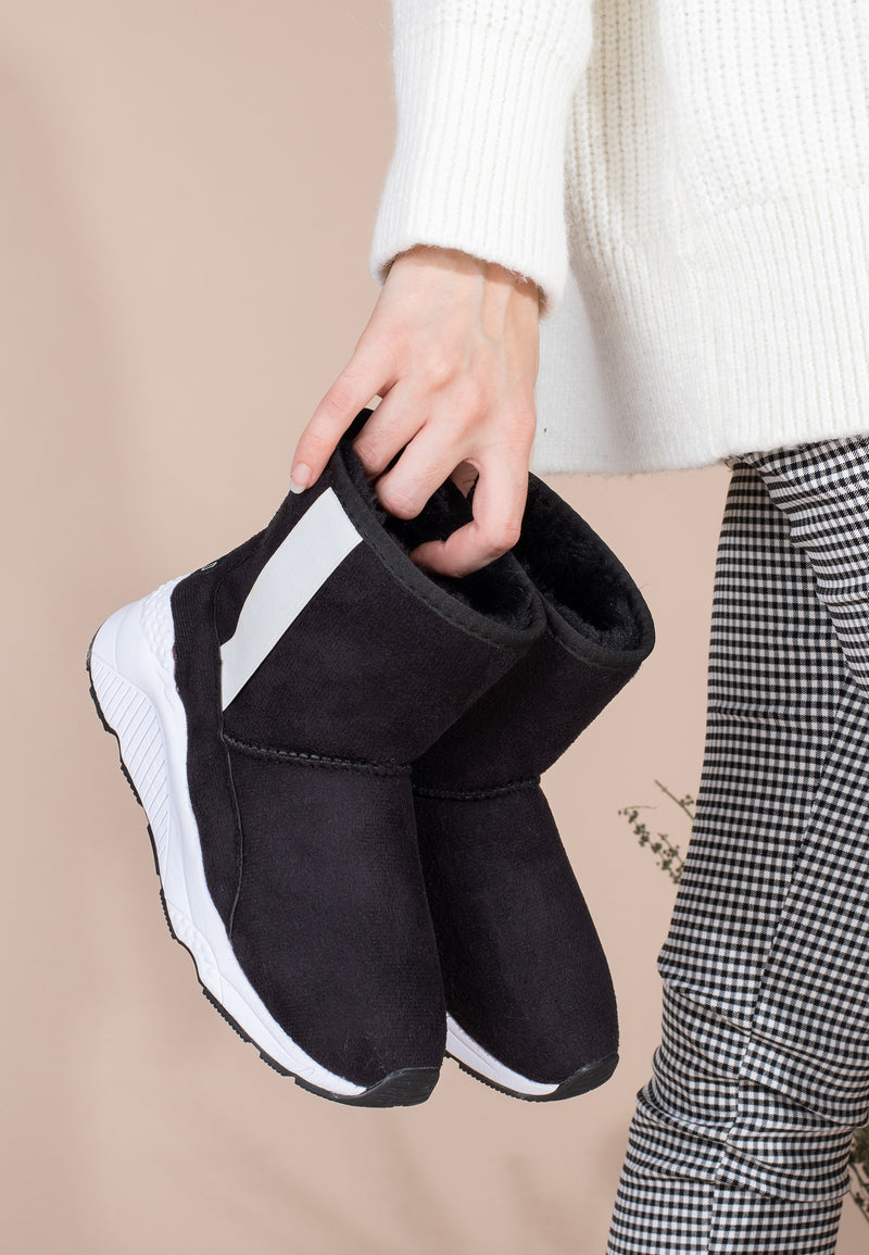 Bottines fourrées Swing noir et blanc tenues à la main, vues de profil devant un pantalon