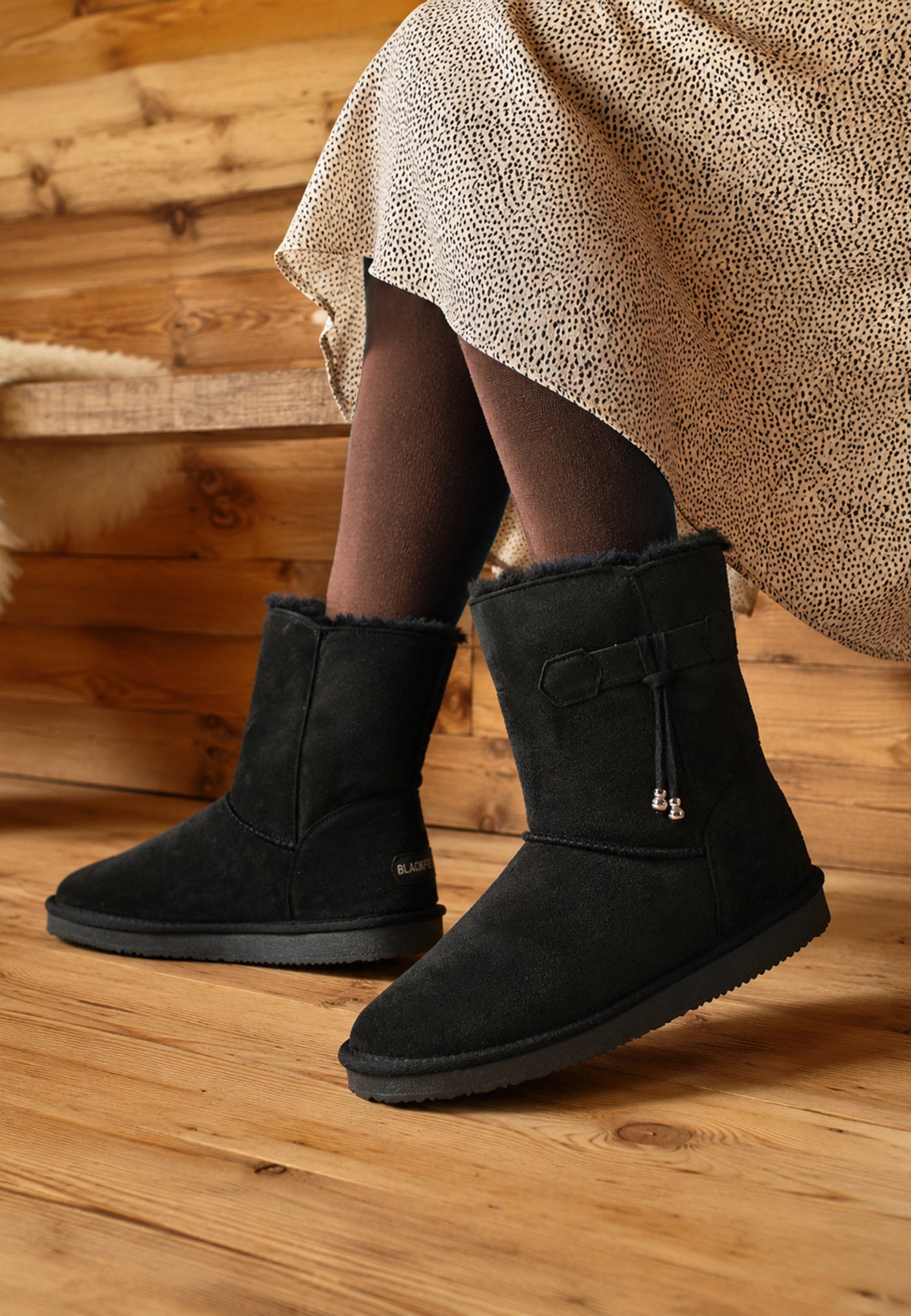 Sur un parquet en bois, bottines fourrées Opinaca noires portées avec une jupe imprimée