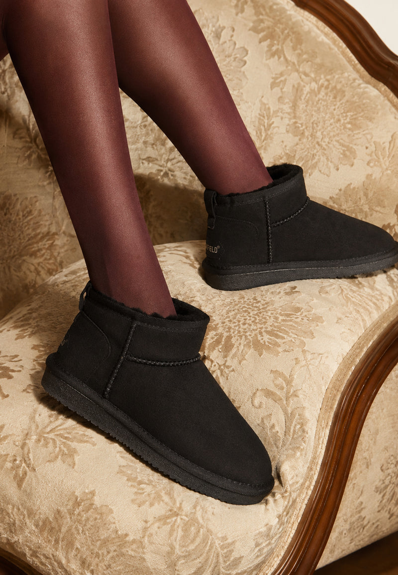 Bottines fourrées Mikela noires portées assise sur un canapé damassé, collants noirs