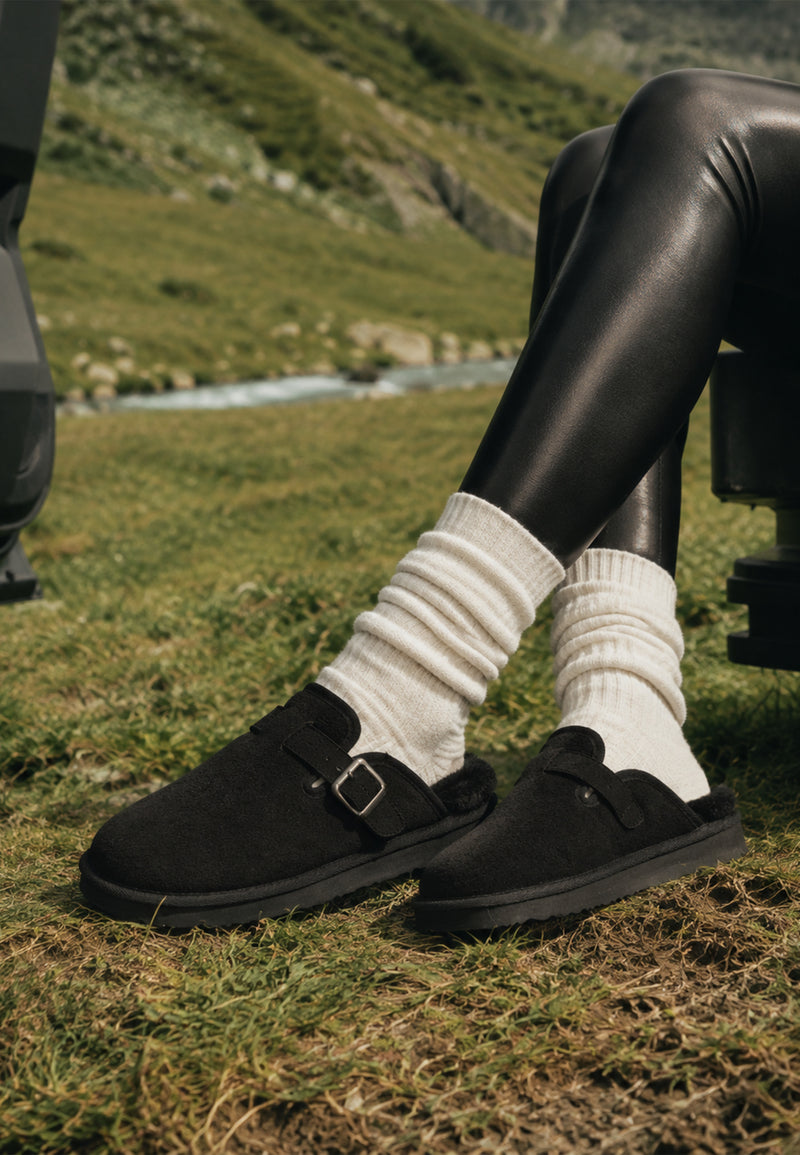 Chaussons fourrés Benoit noirs portés dans l’herbe avec des chaussettes crème